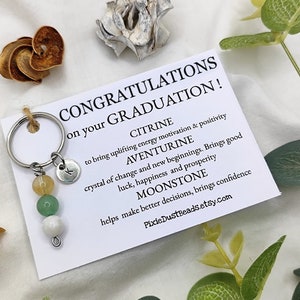 Pode incluir: Um cartão branco com o texto "CONGRATULATIONS on your GRADUATION!" e descrições das propriedades curativas dos cristais de Citrino, Aventurina e Pedra da Lua. O cartão está ao lado de um chaveiro prateado com um amuleto e três contas: uma conta de Citrino amarela, uma conta de Aventurina verde e uma conta de Pedra da Lua branca.