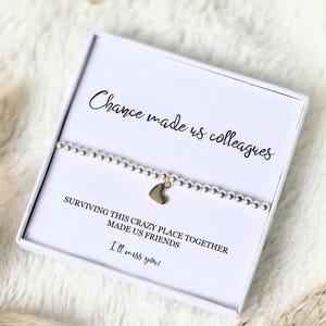 Puede incluir: Una pulsera de cuentas plateadas con un dije de coraz&oacute;n dorado, presentada en una caja de regalo blanca. La caja tiene el texto "Chance made us colleagues, surviving this crazy place together made us friends, I'll miss you!"