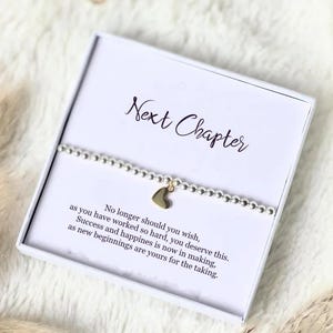 May include: A silver beaded bracelet with a gold heart charm, presented in a white gift box. The box lid reads "Next Chapter" in cursive, with an inspirational message below. A thoughtful gift.