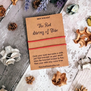 Könnte beinhalten: Eine braune Karte mit dem Text "The Red String of Fate" und Anweisungen für ein Glücksarmband. Die Karte liegt auf einer weißen Oberfläche mit braunen und weißen Dekorationsartikeln.