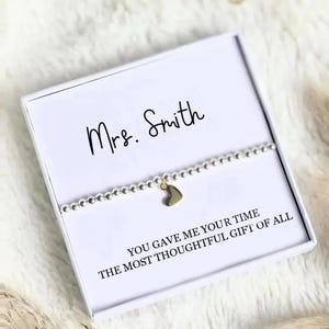 Peut inclure: Un bracelet en perles argentées avec une petite breloque en forme de cœur doré, présenté dans une boîte cadeau carrée blanche. La boîte porte l'inscription "Mrs. Smith" et "YOU GAVE ME YOUR TIME THE MOST THOUGHTFUL GIFT OF ALL" en noir.