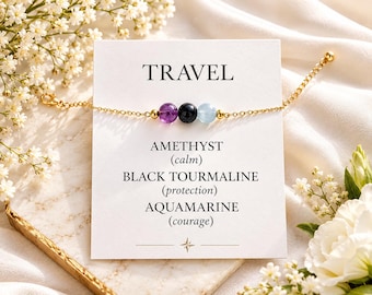 May include: A gold-tone chain bracelet featuring three round beads: a purple amethyst, a black tourmaline, and a light blue aquamarine. The bracelet is displayed on a white card with the text TRAVEL, AMETHYST (calm), BLACK TOURMALINE (protection), and AQUAMARINE (courage).