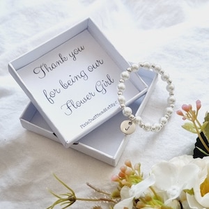 May include: A white box with a message that reads "Thank you for being our Flower Girl" and a white pearl bracelet with a silver charm in the shape of the letter J.