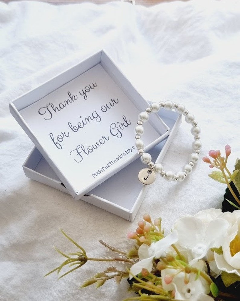 May include: A white box with a message that reads "Thank you for being our Flower Girl" and a white pearl bracelet with a silver charm in the shape of the letter J.