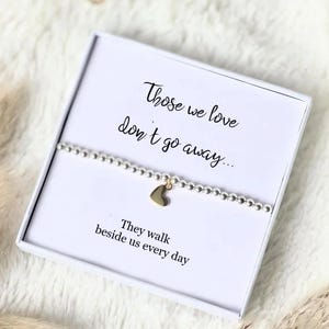 May include: A silver beaded bracelet with a small gold heart charm, presented in a white gift box. The box features the printed text: Those we love don't go away... They walk beside us every day, written in a black script font.