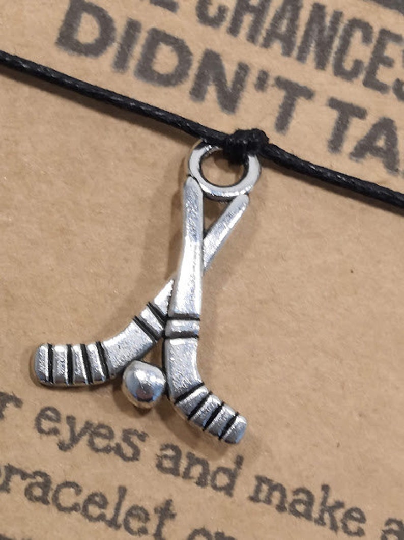 May include: A black cord bracelet with a silver charm of two crossed hockey sticks and a puck. The bracelet has the text "Take Chances You Didn't Take" on the card.