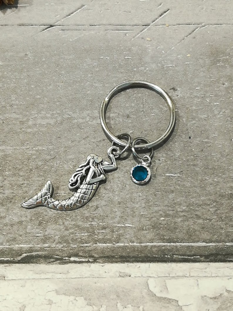 May include: A silver mermaid keychain with a blue gemstone charm attached to a silver key ring.