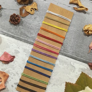 May include: A variety of colored cords are displayed on a brown card. The cords are arranged in rows, with the lightest color at the top and the darkest color at the bottom. The cords are all the same width and are made of a smooth, soft material.
