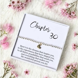 May include: A silver beaded bracelet with a gold heart charm, presented in a white box with the text "Chapter 30". The box is surrounded by pink flowers on a white, fluffy surface. The text on the box reads: "Here's to the dreams that inspire you..."