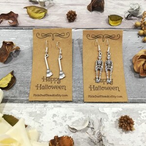 May include: Two pairs of silver Halloween earrings on brown card stock. One pair features a skeleton and the other a broom. The card stock has the text "Happy Halloween" and a website address.