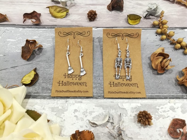 May include: Two pairs of silver Halloween earrings on brown card stock. One pair features a skeleton and the other a broom. The card stock has the text "Happy Halloween" and a website address.
