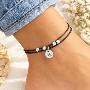 Può includere: Una cavigliera a doppio cordoncino marrone scuro con quattro piccole perline cubiche color argento e un ciondolo centrale rotondo color argento con impressa la lettera maiuscola M in nero.