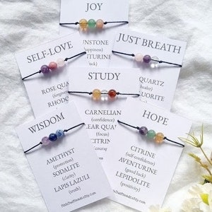 May include: Seven bracelets with different colored gemstones and corresponding words on white cards. The words are: Joy, Self-Love, Wisdom, Just Breath, Study, Hope, and Instone. Each bracelet has a different meaning associated with the gemstones.