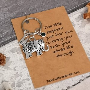 Pu&ograve; includere: Un portachiavi in ​​argento con un ciondolo a forma di elefante, una gemma verde e la lettera "M" appesi a un anello di chiave in argento. Il portachiavi &egrave; su una carta marrone con il testo "This little elephant is just for you to bring you luck your whole life through" e l'indirizzo del sito web "PixieDustReads.etsy.com".
