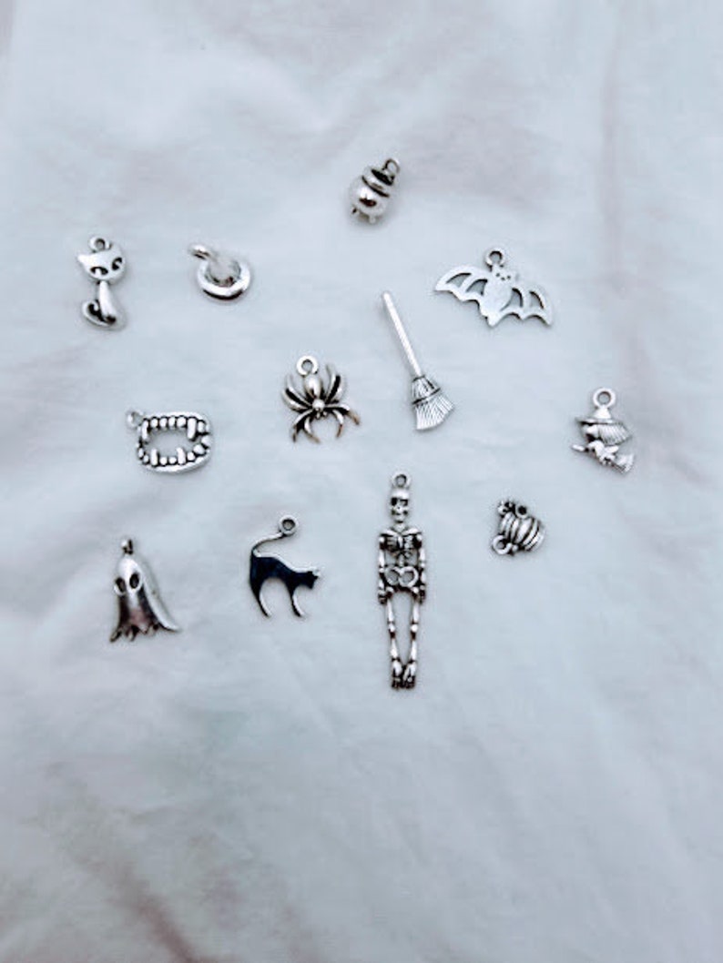 May include: A collection of silver charm pendants with Halloween themes, including a witch's hat, a spider, a ghost, a cat, a skeleton, a bat, a cauldron, a broom, fangs, and a pumpkin.
