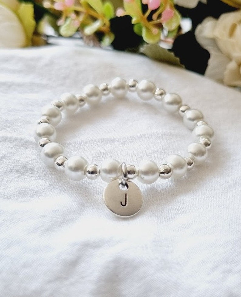 May include: A white pearl bracelet with a silver charm that has the letter "J" on it.