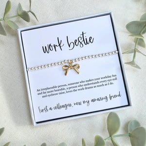 Puede incluir: Una pulsera de cuentas plateadas con un dije de lazo dorado, presentada en una caja de regalo blanca. La caja presenta el texto "work bestie" y un mensaje sobre la amistad. El fondo está adornado con follaje verde.