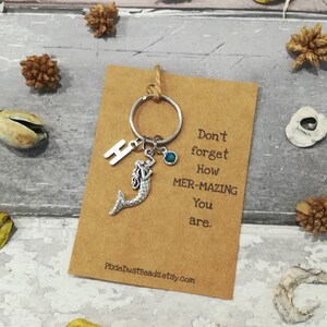 May include: A brown card with the text "Don't forget how mer-mazing you are."  A silver mermaid charm, a silver letter "H" charm, and a blue gemstone are attached to a silver key ring.