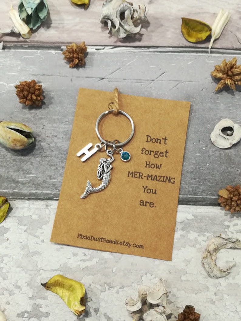 May include: A brown card with the text "Don't forget how mer-mazing you are."  A silver mermaid charm, a silver letter "H" charm, and a blue gemstone are attached to a silver key ring.
