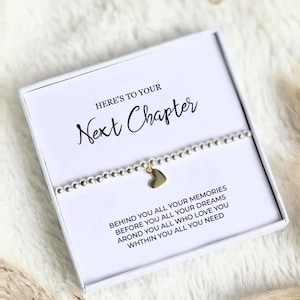 May include: A white gift box with the text "HERE'S TO YOUR Next Chapter" and a silver beaded bracelet with a gold heart charm. The box also includes the text "BEHIND YOU ALL YOUR MEMORIES BEFORE YOU ALL YOUR DREAMS AROND YOU ALL WHO LOVE YOU WHTHIN YOU ALL YOU NEED."
