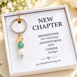 May include: A silver-toned metal keyring featuring three spherical beads in yellow, green, and white, presented in a white box with text reading NEW CHAPTER, MOONSTONE (new beginnings), ADVENTURINE (luck), CITRINE (energy), and pixiedustbeads.etsy.com.