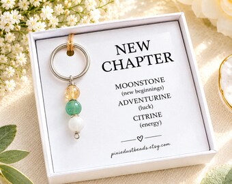 May include: A silver-toned metal keychain featuring three spherical beads in yellow, green, and white, presented in a white box with text reading NEW CHAPTER, MOONSTONE (new beginnings), ADVENTURINE (luck), CITRINE (energy), and pixiedustbeads.etsy.com.