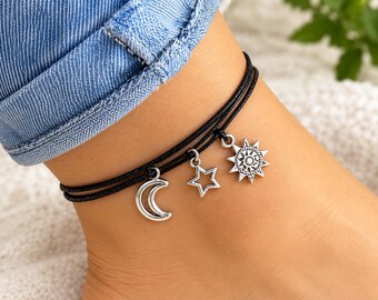 May include: A double-strand black cord anklet featuring three silver-toned metal charms: a crescent moon, an open-work star, and a detailed sun with radiating points. The charms are evenly spaced along the thin, dark, braided-style cord.