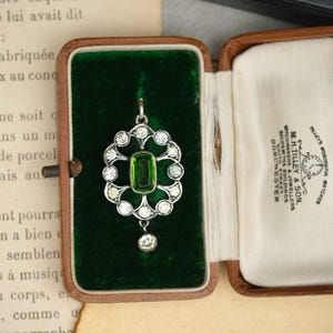 May include: A silver-toned pendant featuring a central rectangular green stone surrounded by a circular arrangement of small clear stones, with a single clear stone drop at the bottom, presented inside a brown velvet-lined jewelry box.