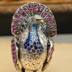 May include: A silver peacock figurine featuring a vibrant blue enameled neck and chest with textured feather detailing. The fan-shaped tail and wings are adorned with numerous small, round, deep pink gemstones set into the metallic silver surface.