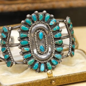May include: A silver cuff bracelet featuring a large oval centerpiece surrounded by small, teardrop-shaped turquoise stones. The central design includes two curved silver elements and two small turquoise stones set in twisted silver wire.