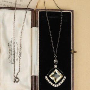 May include: A dark metal necklace featuring a square-cut, pale yellow gemstone pendant suspended within a diamond-shaped frame, accented by a curved row of small clear stones at the base, presented in a rectangular black-lined jewelry box.