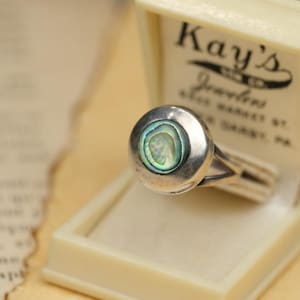 May include: A silver ring featuring a circular setting with an iridescent abalone shell inlay. The ring is displayed inside a cream-colored jewelry box with the text Kay's Jewelers, 69th Market St., Upper Darby, PA printed on the interior lid.
