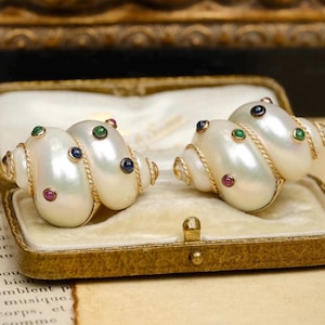 May include: A pair of shell-shaped earrings featuring a lustrous white finish with gold-tone twisted rope accents. Each earring is embellished with small, round, multi-colored gemstones in shades of green, blue, and red set into the surface.