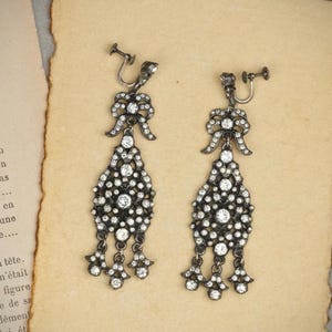 May include: A pair of dark metal dangle earrings featuring a bow-shaped top section and a larger, ornate, teardrop-shaped lower section, both encrusted with numerous small, clear, sparkling stones. The earrings are secured with screw-back fasteners.