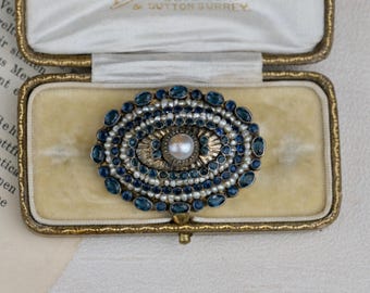 May include: An oval brooch with a central pearl, surrounded by concentric circles of small pearls and blue gemstones. The brooch is set in a gold-toned metal and displayed in a vintage jewelry box with a cream-colored interior.