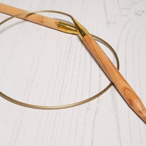 Könnte beinhalten: Ein Satz runder Stricknadeln aus Holz mit einem goldfarbenen Metallkabel. Die Nadeln sind spitz und haben eine natürliche Holzoptik.