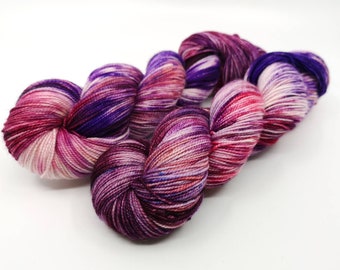 May include: A skein of hand-dyed yarn in shades of pink, purple, and white. The yarn is a variegated colorway with a speckled appearance.