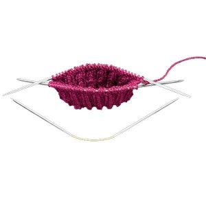 Op de afbeelding: Een close-up van een breiproject in uitvoering, met een diep magenta gebreid stuk met een ribbelpatroon. Zilveren breinaalden zijn in het werk gestoken en een draad in magenta is bevestigd. De naalden zijn rond het gebreide item geplaatst.
