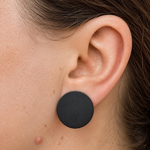 May include: A matte black circular stud earring featuring a flat, smooth surface. The earring is designed as a simple, solid disc shape that sits flush against the earlobe, providing a clean and understated appearance for everyday wear.