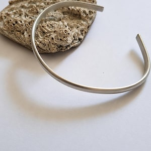 Op de afbeelding: Een zilverkleurige metalen armband met een gladde, matte afwerking en een open ontwerp. De armband is gebogen in een cirkelvorm en heeft een eenvoudige, onversierde band die over de gehele lengte een constante dikte behoudt.