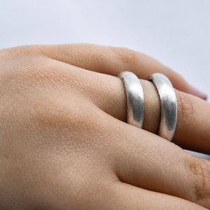 May include: Two silver-coloured rings with a brushed, matte texture. The bands have a rounded, smooth profile and a consistent width, featuring a subtle metallic sheen across the surface of the metal, worn on two adjacent fingers.
