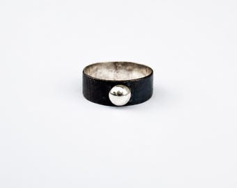Può includere: Un anello a fascia larga con una finitura esterna nera ossidata e scura, caratterizzato da una singola sfera color argento lucido posizionata al centro. L'interno dell'anello presenta una finitura argento metallizzato brillante, in contrasto con l'esterno.