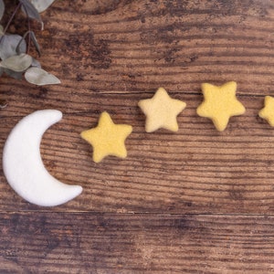 Op de afbeelding: Een maan- en sterrenset van vilt in wit en geel. De halve maan is wit en vijf gele sterren van verschillende grootte zijn in een rij gerangschikt. De objecten staan op een houten oppervlak, met een takje groen linksboven.