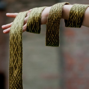 May include: A woven belt in shades of olive green and gold is held up in a hand. The belt features a geometric diamond pattern and a black metal D-ring. The background is a blurred brick wall and greenery.
