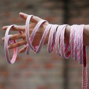 May include: A hand wrapped in a long, white and red woven cord with a geometric pattern. The cord is tied around the wrist and hangs down, creating a decorative effect.