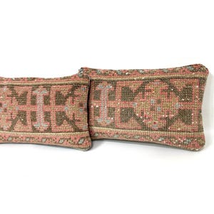 Puede incluir: Dos cojines rectangulares con un patr&oacute;n textil tejido en tonos apagados de rosa polvoriento, verde oliva y crema. La tela muestra un motivo geom&eacute;trico repetitivo con fibras texturizadas y gruesas y costuras visibles en los bordes.