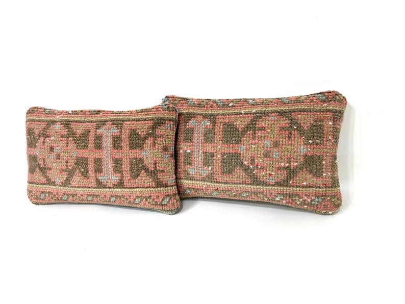 Puede incluir: Dos cojines rectangulares con un patr&oacute;n textil tejido en tonos apagados de rosa polvoriento, verde oliva y crema. La tela muestra un motivo geom&eacute;trico repetitivo con fibras texturizadas y gruesas y costuras visibles en los bordes.