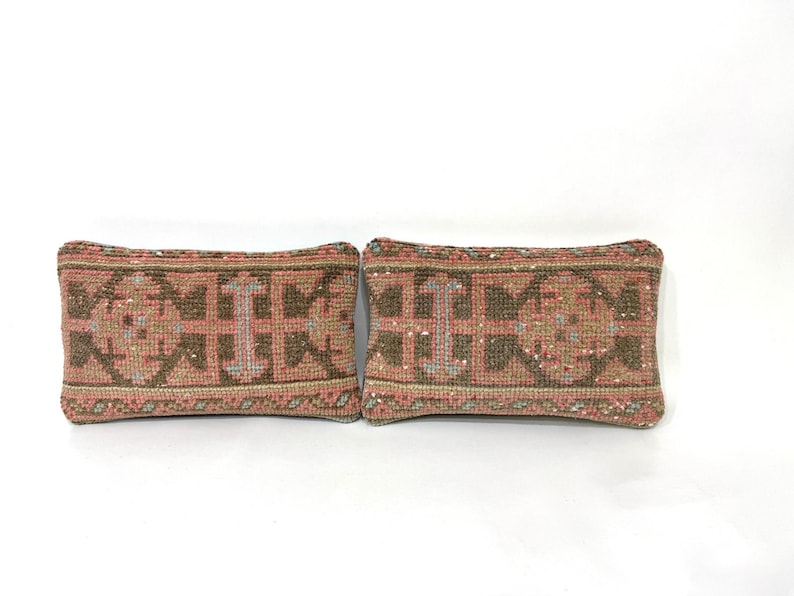 Puede incluir: Dos cojines rectangulares con un patr&oacute;n textil tejido en tonos rosa empolvado, marr&oacute;n y crema. La tela muestra un motivo geom&eacute;trico repetitivo con fibras texturizadas y gruesas, adem&aacute;s de bordes acabados, presentando un dise&ntilde;o uniforme.