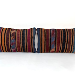 May include: Two rectangular throw pillows with a striped pattern in shades of orange, burgundy, black, and blue. The pillows have a woven texture and a blue border. The design is symmetrical.