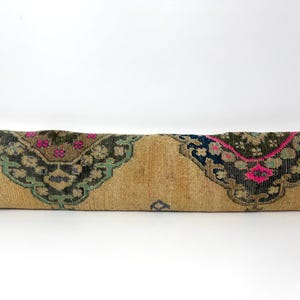 Peut inclure: Un long coussin rectangulaire avec un fond beige et des extrémités décoratives à motifs. Les extrémités présentent un motif vert foncé et marron avec des accents roses et bleus. Le coussin est probablement fait d'une matière tissée.